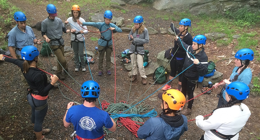 a group of people wearing helmets stand in a circle and are connected by a tangle of ropes at the center. 